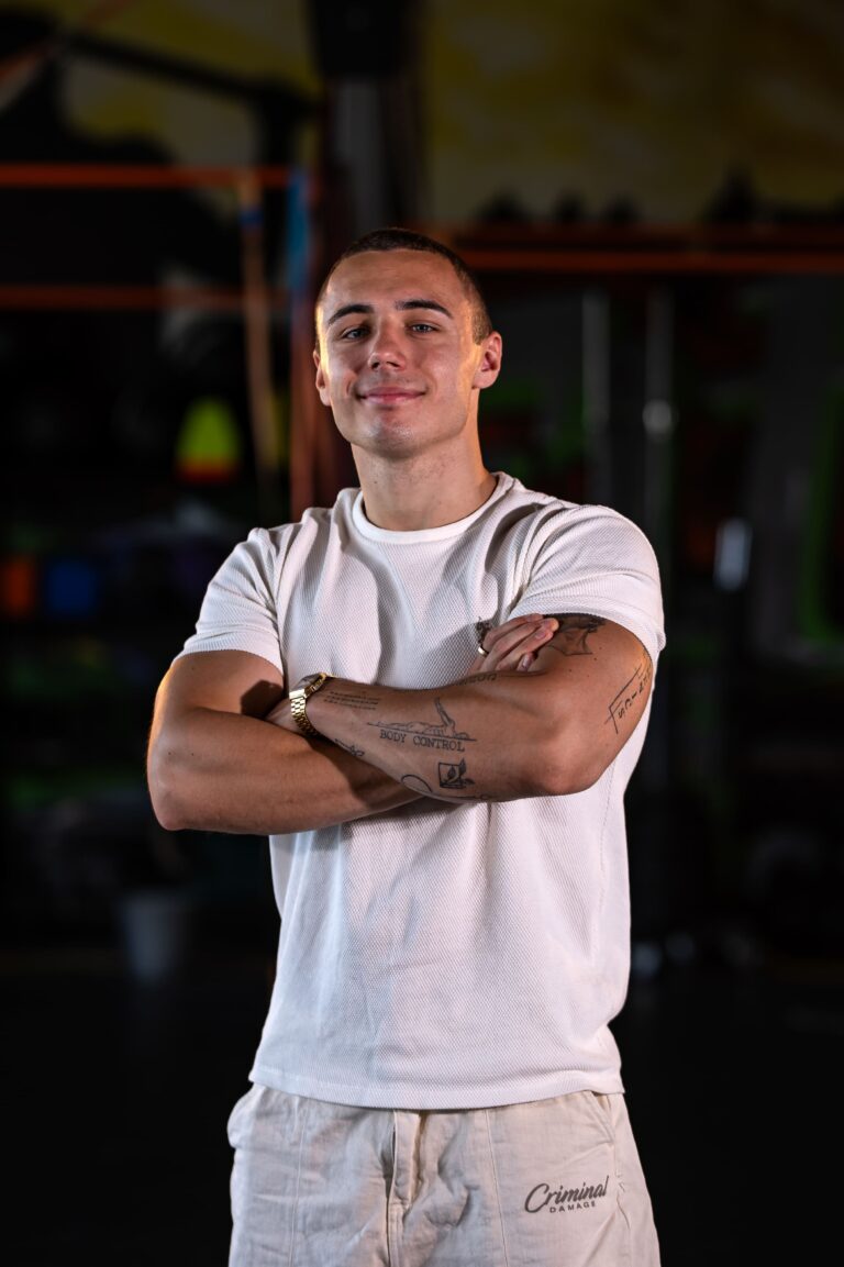 Young man with short hair in a white T-shirt, arms crossed, standing in a gym with equipment in the blurred background.