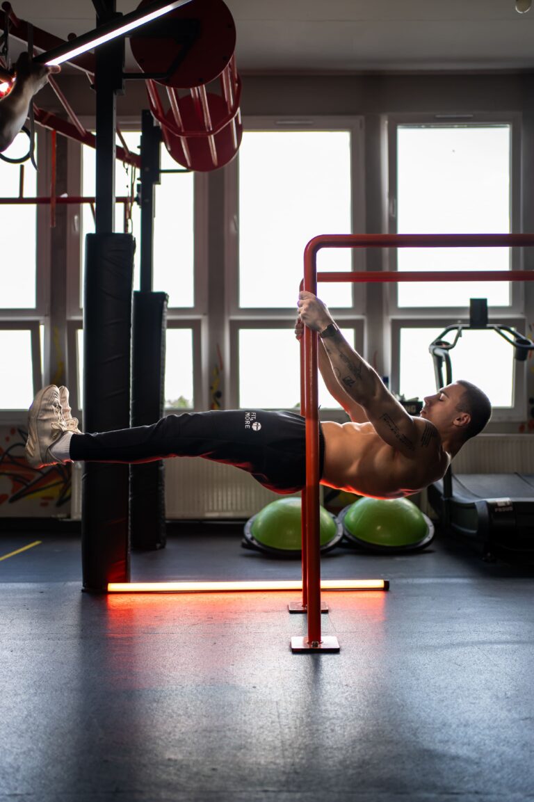 Shirtless man performing leg raises on red parallel bars in a gym, with large windows in the background.
