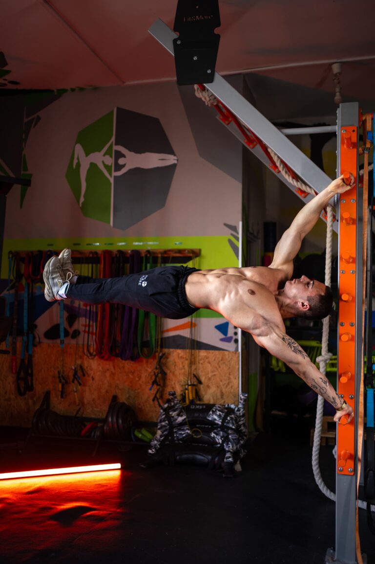 Man performing a hanging leg raise on an orange gym rack in a colorful gym, torso exposed and legs extended behind him.