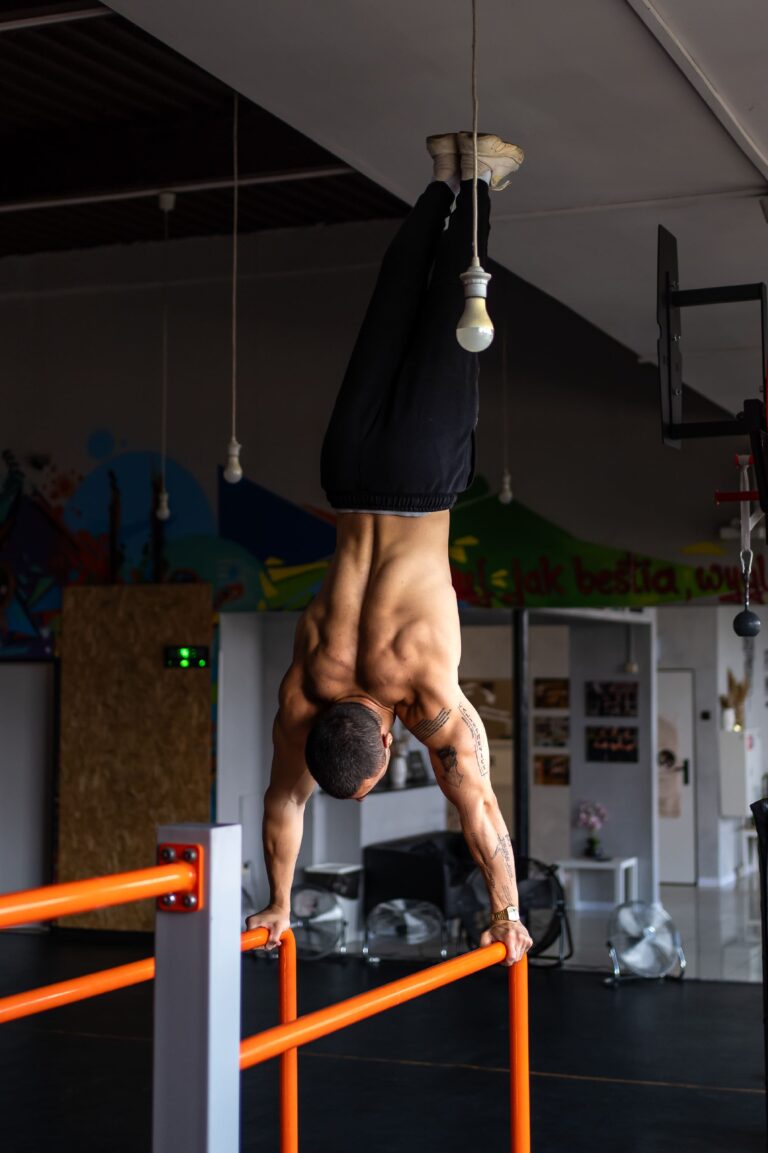 Man in a gym hanging upside down from orange parallel bars, gripping with hands, torso extended and head facing downward during a workout session.