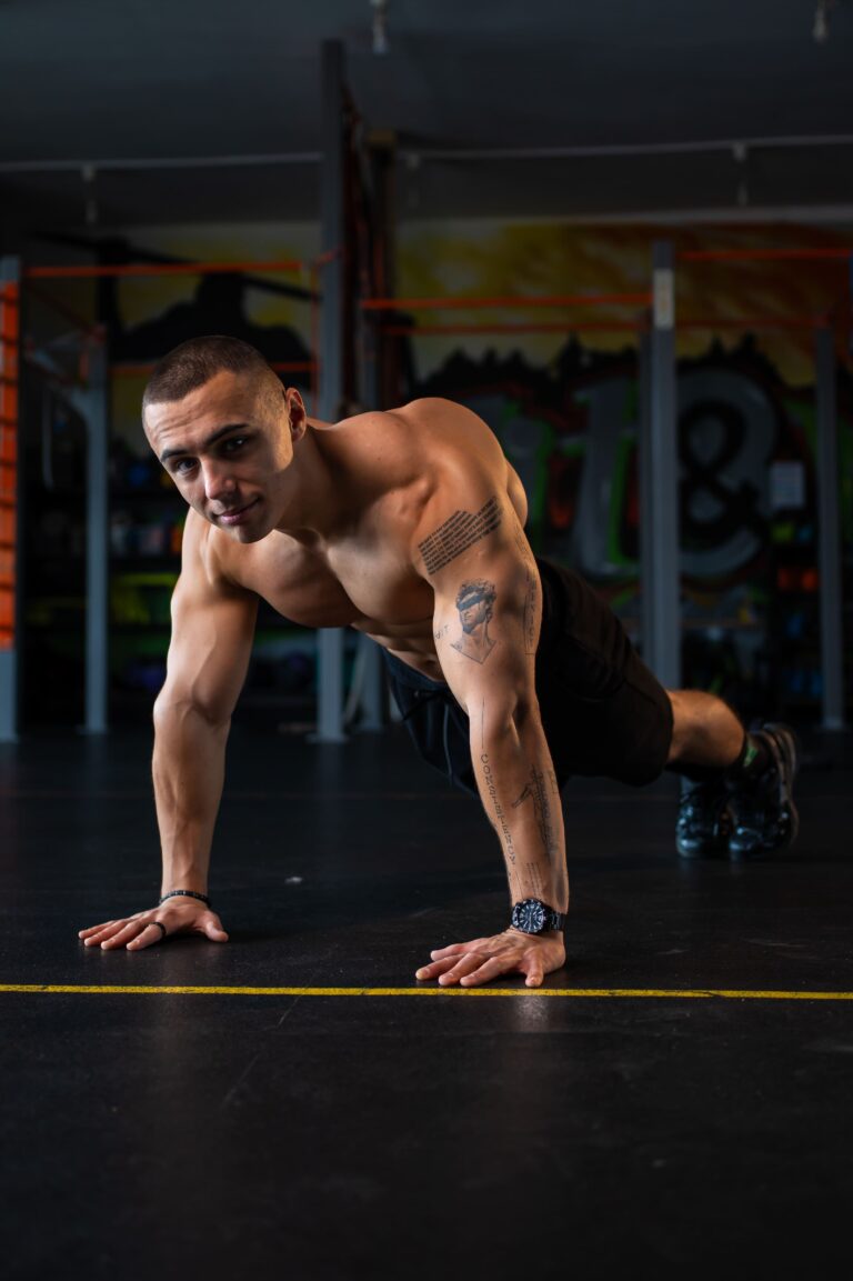 Young man performing a push-up in a gym, shirtless, tattoos on his left arm, focused ahead.