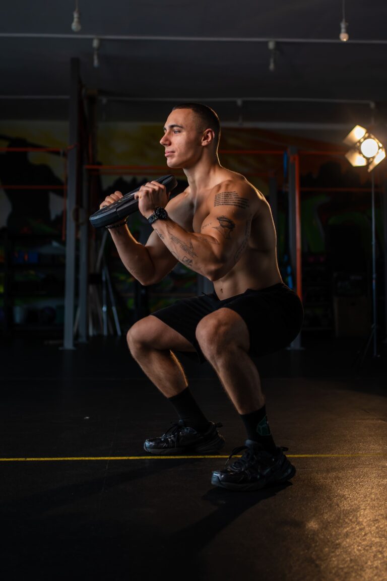 A shirtless man in a gym performs a squat holding a weight plate across his chest, tattoos visible on his arm, focused forward as lighting highlights him.
