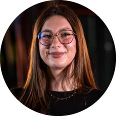 Portrait of a woman with shoulder-length brown hair, round glasses, and a subtle smile, wearing a dark top against a blurred background.