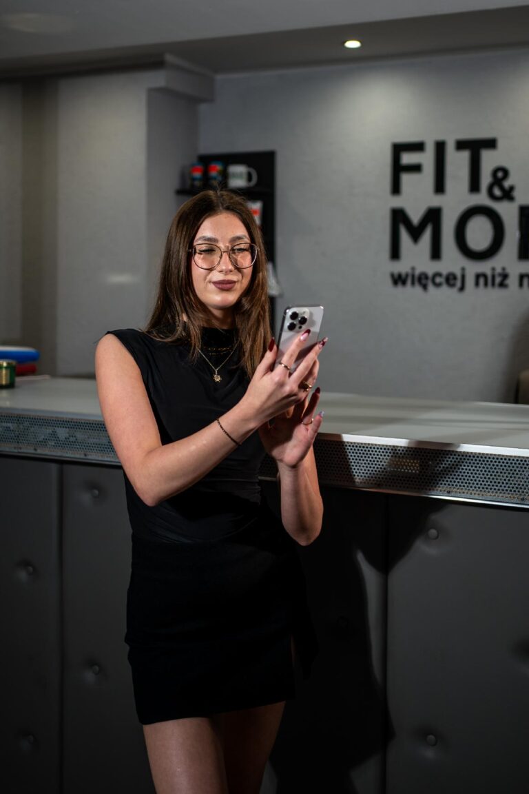 Woman in a black sleeveless dress using a smartphone at a metallic counter in a modern gym/fitness space. Behind her, a wall with large text reads 'FIT & MO...'.