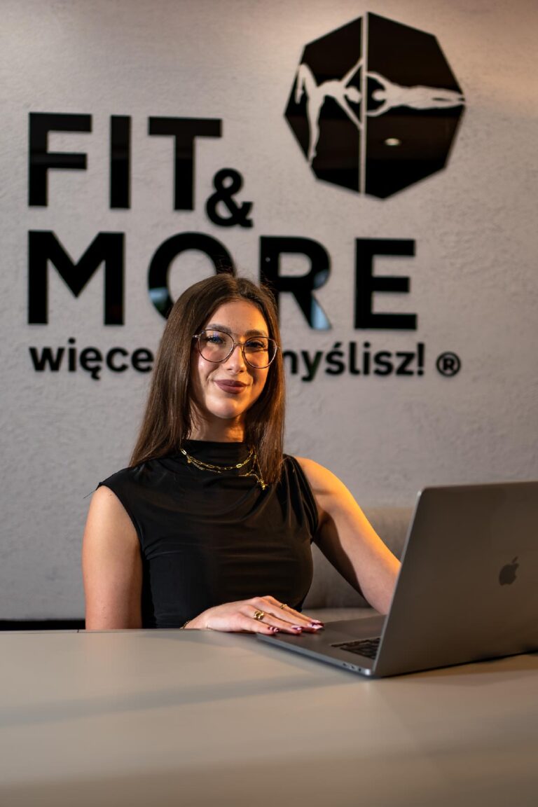 Woman with glasses sits at a desk, smiling, working on a silver laptop in front of a wall with a large 'FIT & MORE' logo.