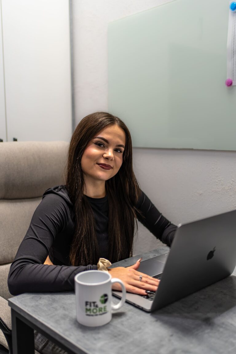 Smiling woman in a black top sits at a gray desk with a laptop, coffee mug, and a glass board behind her.