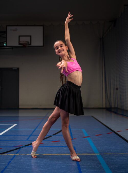 Young female dancer in a pink sports top and black skirt mid-dance in a gym, one arm raised gracefully and smiling.