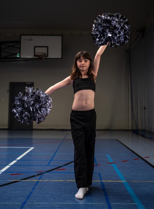 Young girl in a black sports top and pants cheerleading with raised arms and silver pom-poms in a gym.