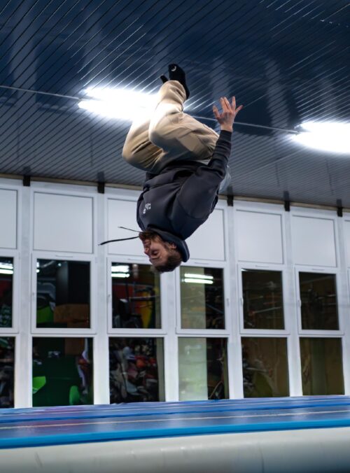 Person in a dark hoodie upside down, mid-flip on a trampoline in an indoor gym.