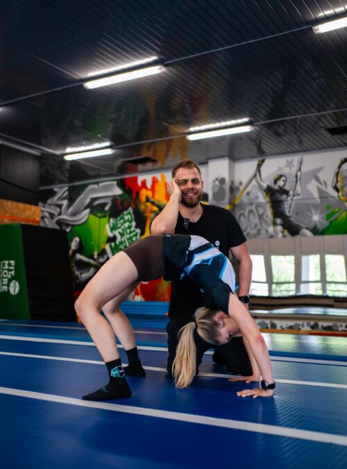 Man in a black shirt smiles while assisting a woman in a backbend during partner yoga in a graffiti-filled gym with blue mats.