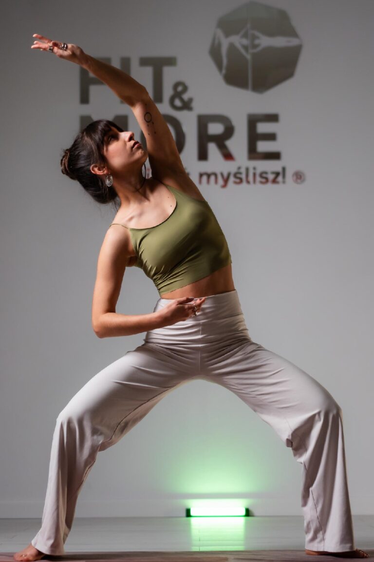 Woman practicing yoga in a studio, arms raised overhead in a wide stance, wearing an olive green sports top and beige pants, green light behind." (concise)