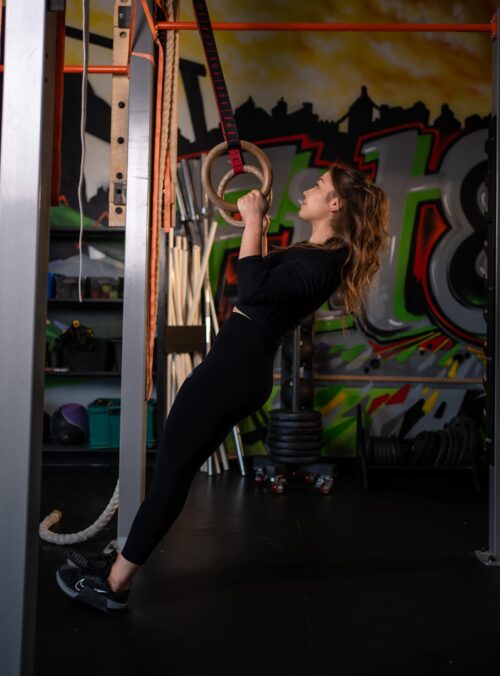 Woman in a black outfit performing a gym ring exercise, gripping wooden rings suspended by straps in a graffiti-filled gym.