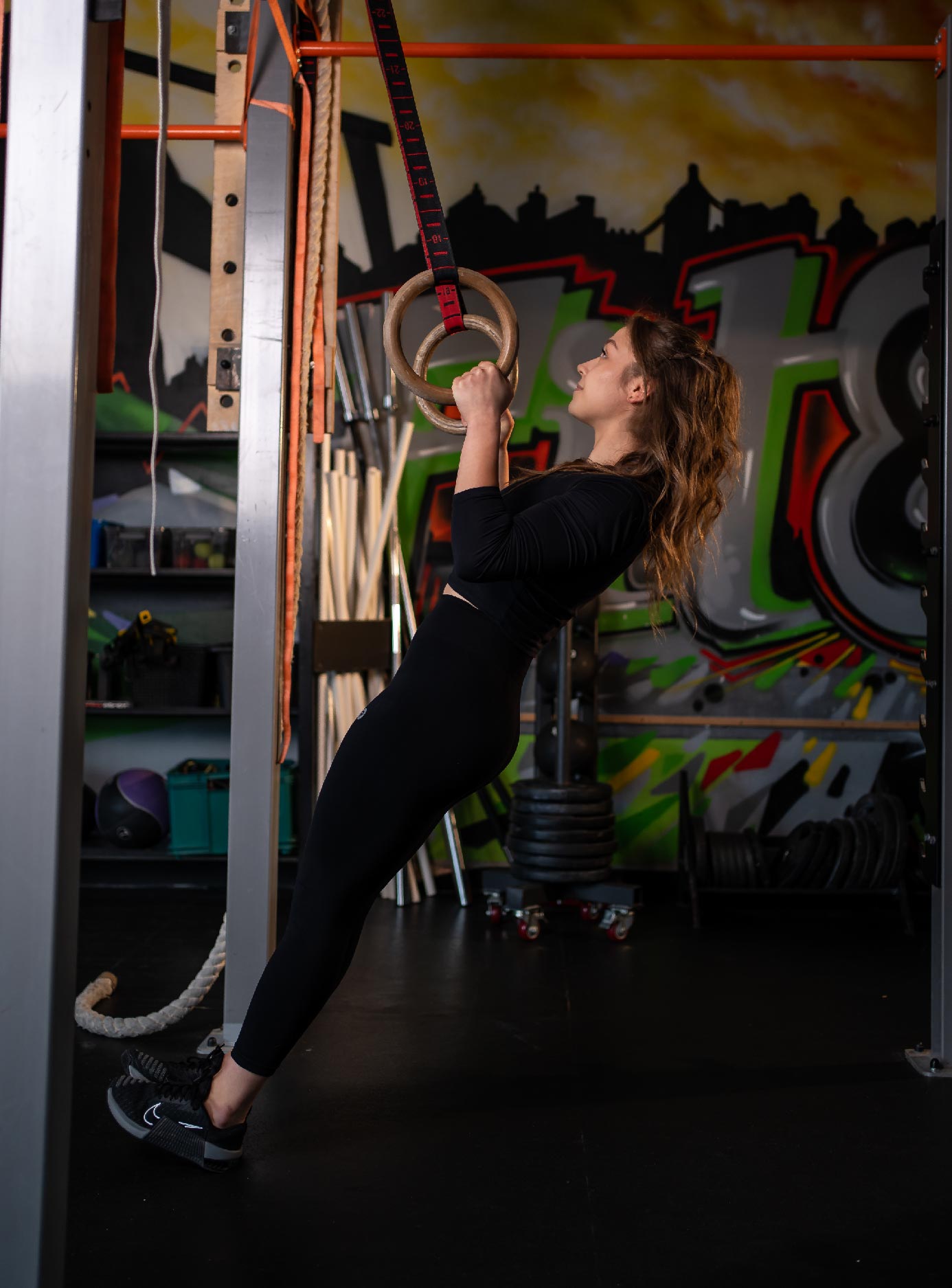 Woman in a black outfit performing a gym ring exercise, gripping wooden rings suspended by straps in a graffiti-filled gym.