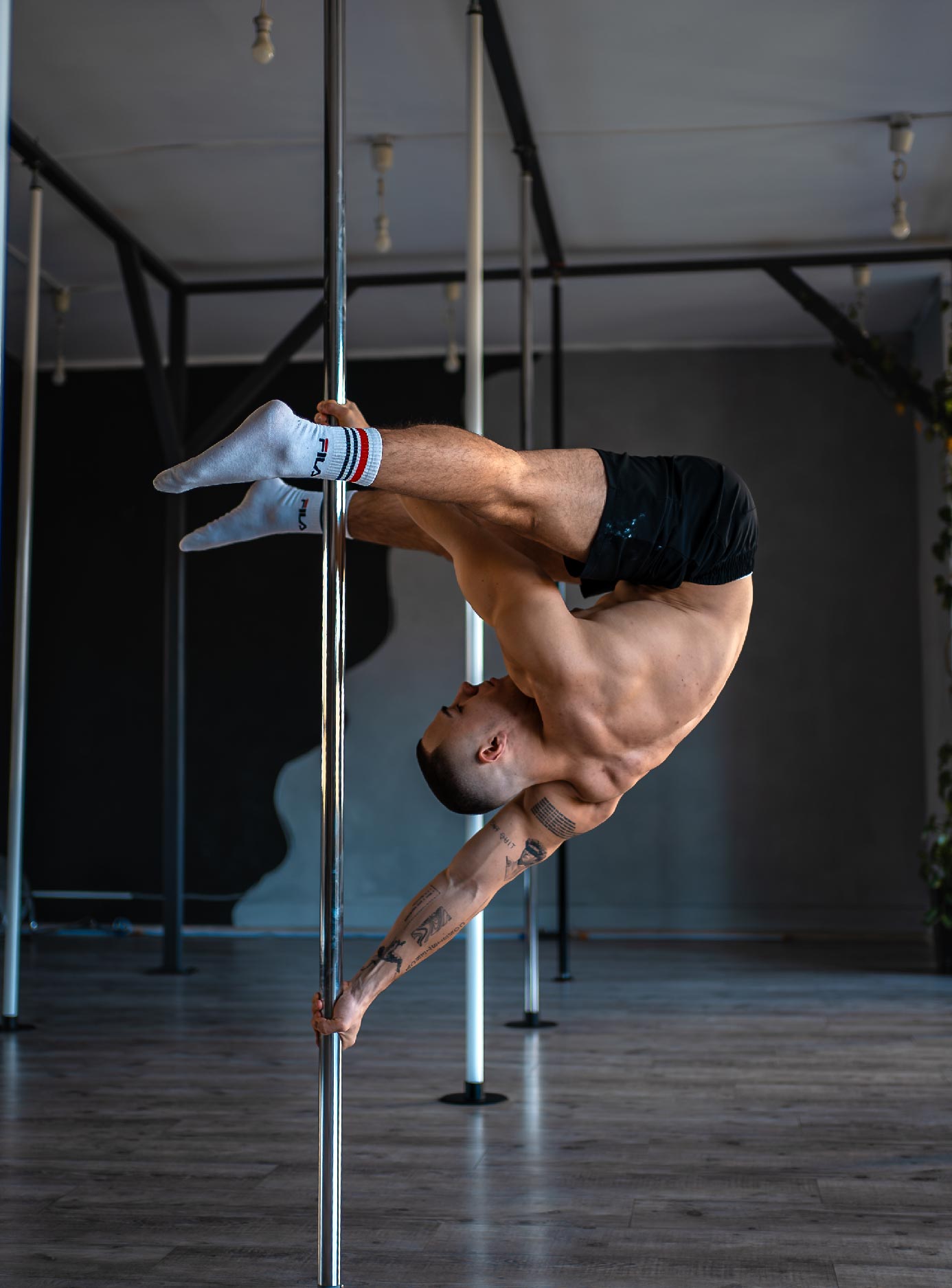 Man performing an inverted pole dance move, gripping a vertical pole with legs wrapped around it in a gym studio.