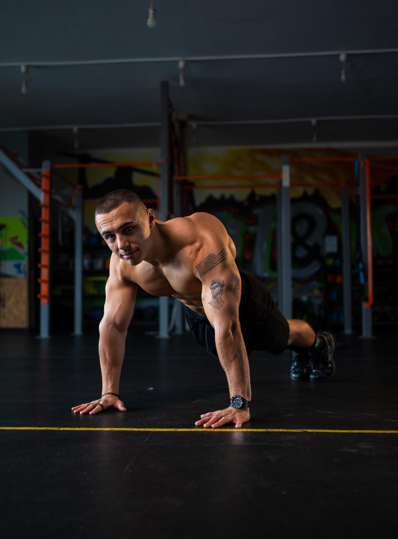 A shirtless man with tattoos performs a push-up in a gym, balancing on hands and toes.