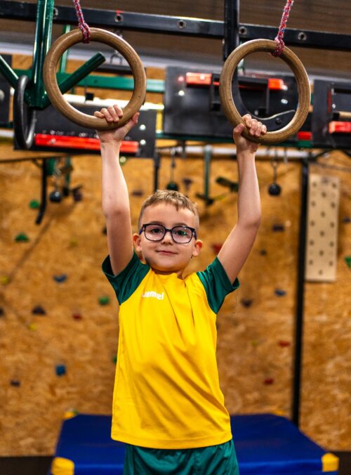 Boy in a yellow and green uniform grips wooden gym rings overhead in an indoor gymnasium door frame setup, looking at the camera.