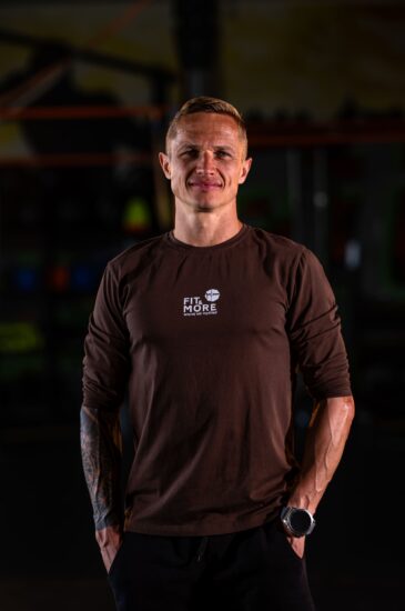 Portrait of a smiling man in a brown shirt with 'FIT & MORE' logo in a gym setting, arms at his sides, looking at the camera.