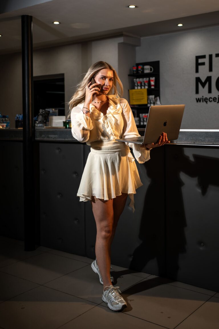 Woman in a white dress speaks on a cellphone while holding a silver laptop at a modern counter.