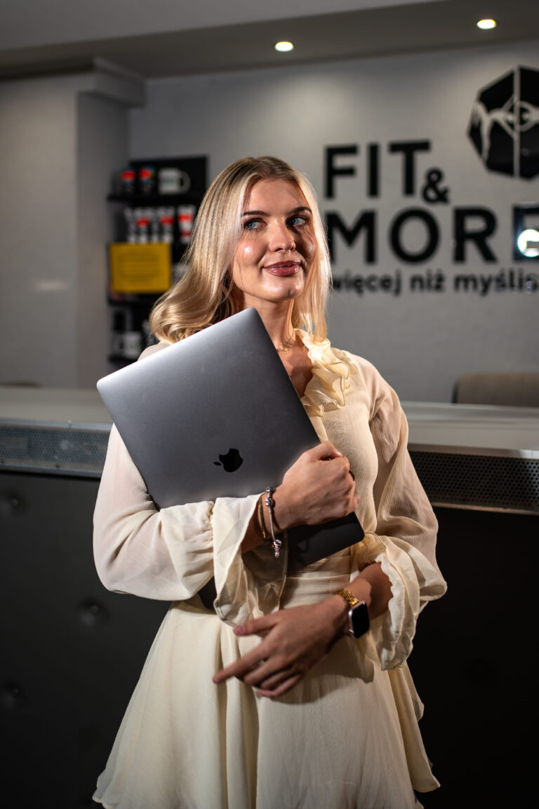 Woman in a cream dress holds a silver laptop, standing in a modern office with bold "FIT & MORE" signage in the background.