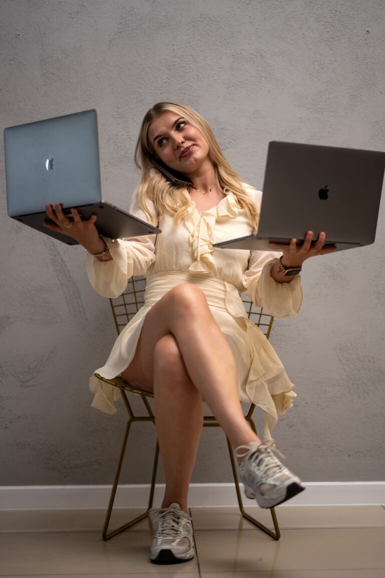 Woman in a cream dress sits on a wire chair, juggling a laptop in each hand while talking on the phone between them.
