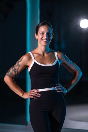 Smiling woman in black-and-white athletic wear stands with hands on hips in a dark studio with blue lighting.