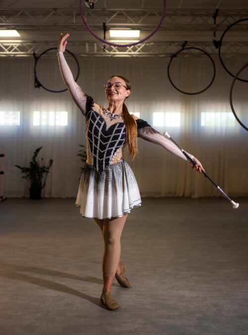 Mazoretki_01_zajecia-fitmore-rybnik_ - Fit&More Rhythmic gymnast in a sparkly leotard raises a purple hoop while smiling in a practice hall with other hoops in the background.