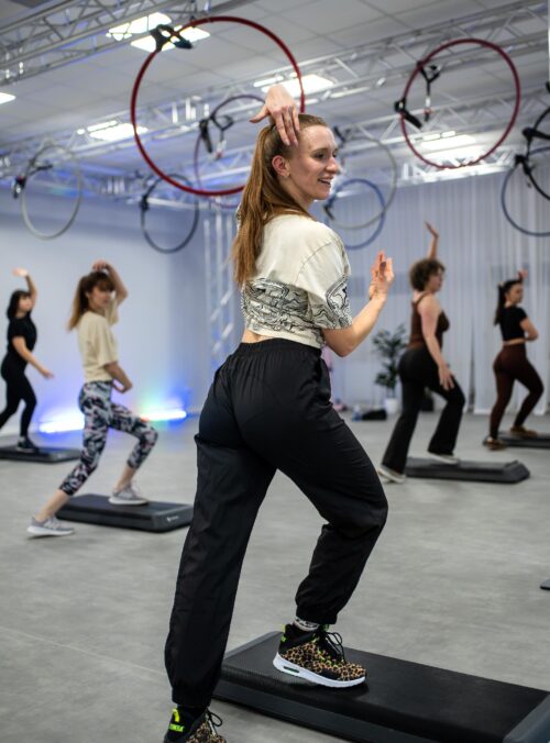Instructor leading a step aerobics class in a studio with hanging hoops, as participants follow the routine on steps.