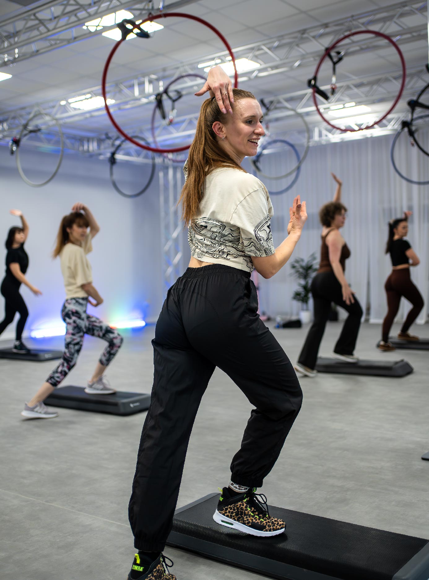 Step-Dance_01_zajecia-fitmore-rybnik_ - Fit&More Instructor leading a step aerobics class in a studio with hanging hoops, as participants follow the routine on steps.