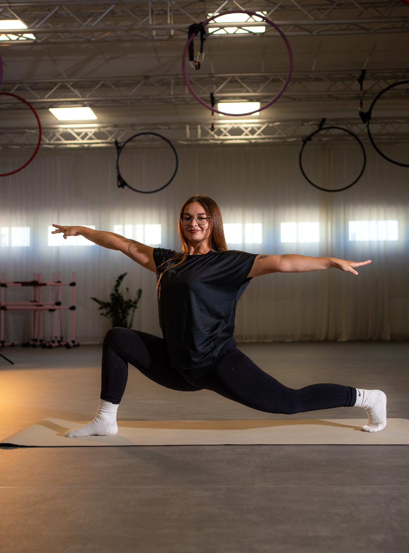Stretching-Kids_01_zajecia-fitmore-rybnik_ - Fit&More Woman in a black workout outfit performs a low lunge split on a mat in a gym, arms extended sideways.