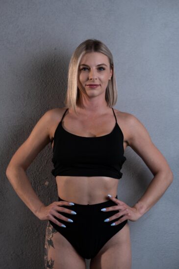 Woman posing in a black bikini against a gray textured wall, hands on hips.