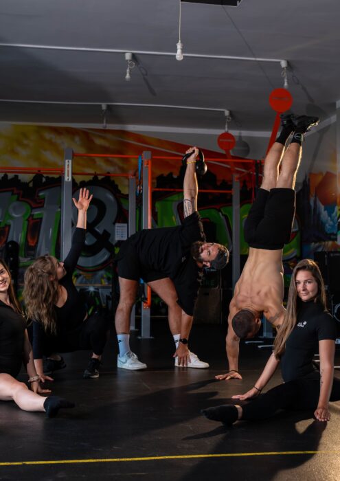 Group of young adults performing a gymnastic routine indoors, balancing hands and legs in the air amid colorful graffiti walls and gym equipment behind them.