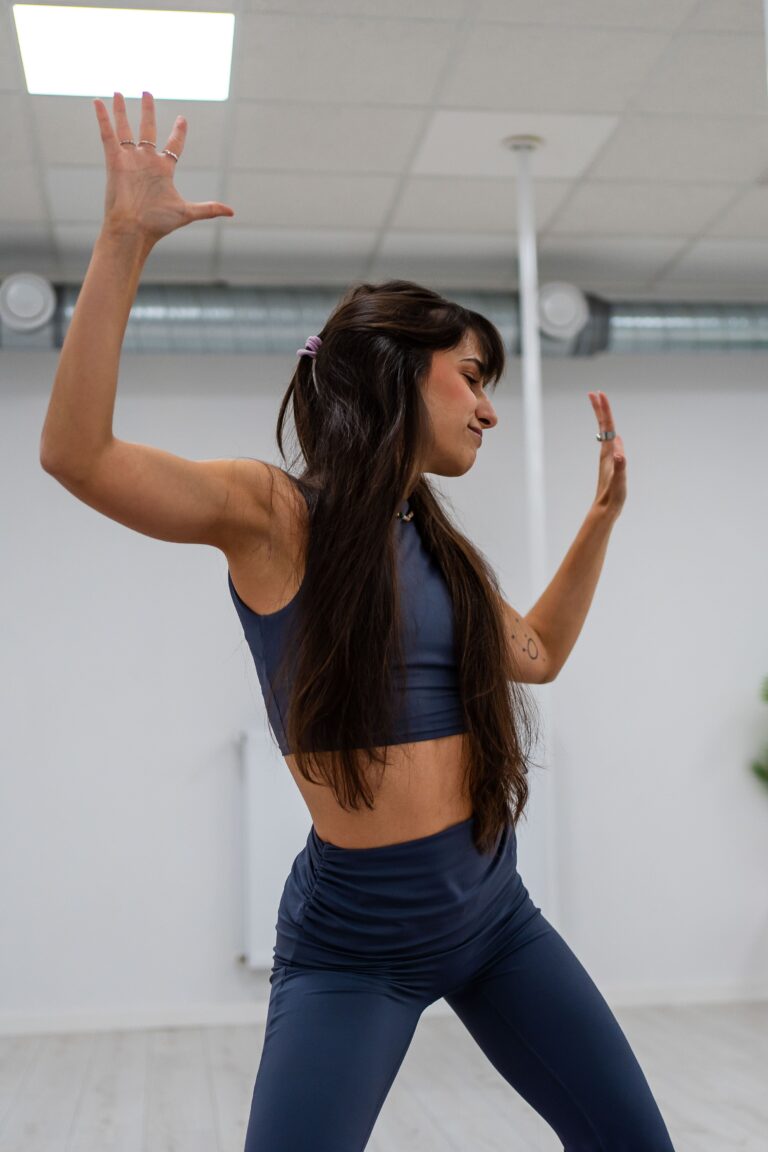 Woman in navy athletic wear raises both arms in a dynamic workout pose in a bright indoor studio.
