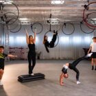 Fitmore 17 lut-20-3 - Fit&More Group of young athletes practicing aerial hoop and balance tricks in a bright gym studio with truss overheads and hoops hanging from above.