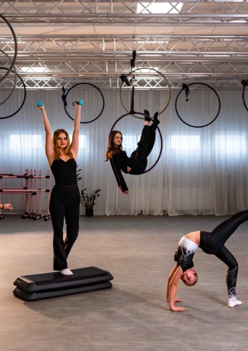 Fitmore 17 lut-20-3 - Fit&More Group of young athletes practicing aerial hoop and balance tricks in a bright gym studio with truss overheads and hoops hanging from above.