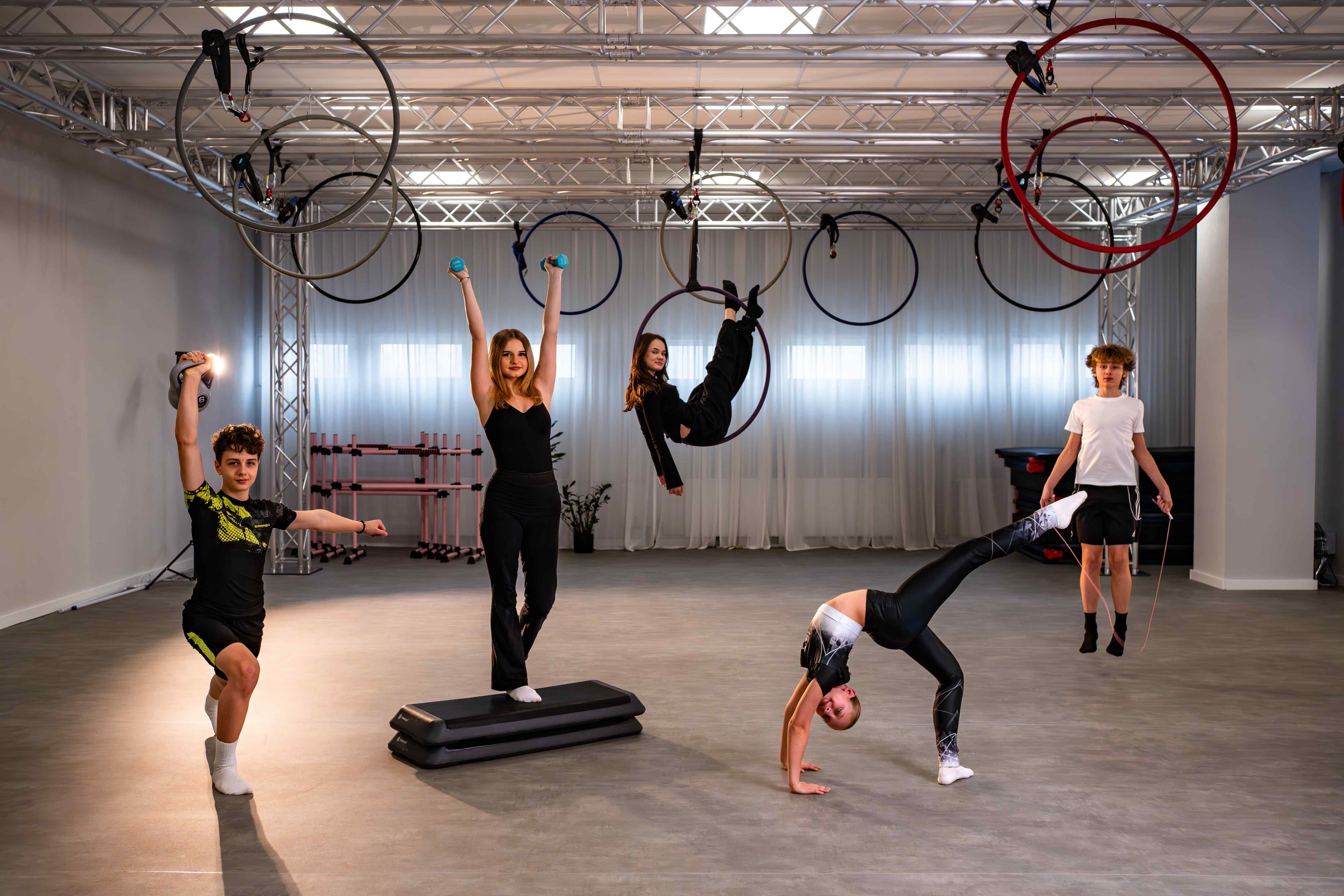 Group of young athletes practicing aerial hoop and balance tricks in a bright gym studio with truss overheads and hoops hanging from above.