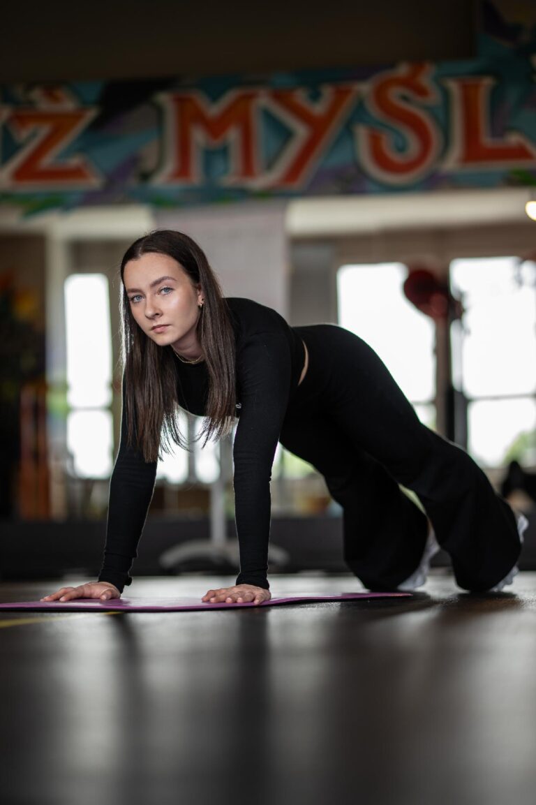 Woman in black athletic wear performs a plank on a pink yoga mat in a gym with a colorful mural in the background.