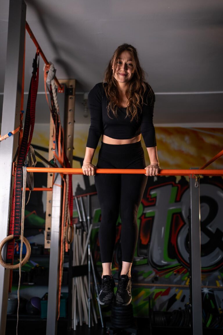 Young woman in a black workout outfit hanging from an orange bar in a gym, smiling at the camera.