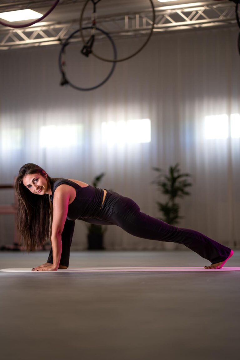 Woman in black athletic wear performs a plank exercise on a pink mat in a bright gym studio.