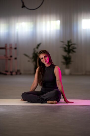 Woman seated cross-legged on a white mat in a bright gym studio, pink light highlighting her side.