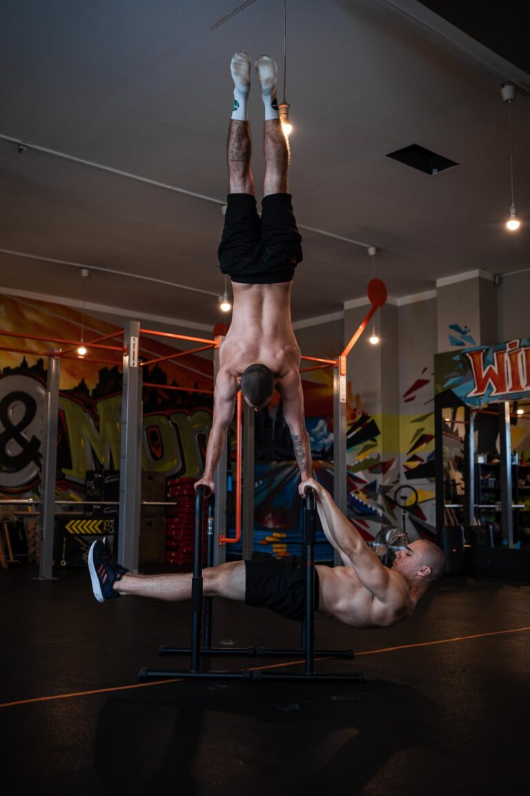Street-workout-skills_zajecia-fitmore-rybnik - Fit&More Two men in a gym: one is upside down, gripping parallel bars, while the other lies on the floor and holds his ankles for support.