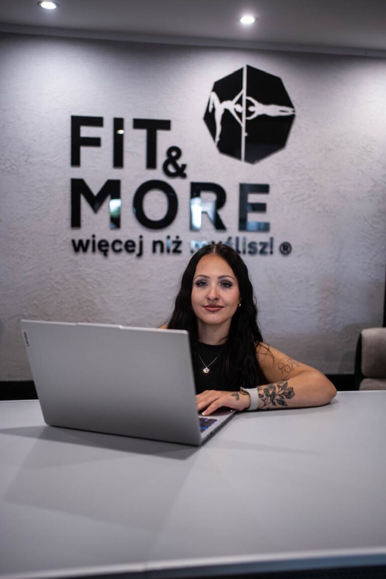 Woman with dark hair seated at a desk, typing on a silver laptop in front of a textured wall with 'FIT & MORE' logo behind her.