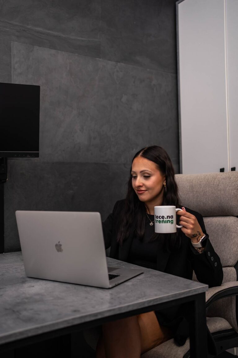 Woman seated at a desk with a silver laptop, holding a white mug, in a modern office setting.
