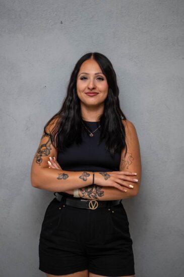 Woman with long dark hair and tattoos, wearing a black sleeveless top and black shorts, arms crossed against a gray wall.