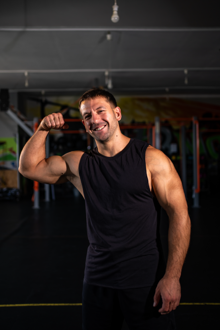 Smiling man in a black sleeveless shirt flexing his bicep in a gym environment, facing the camera.