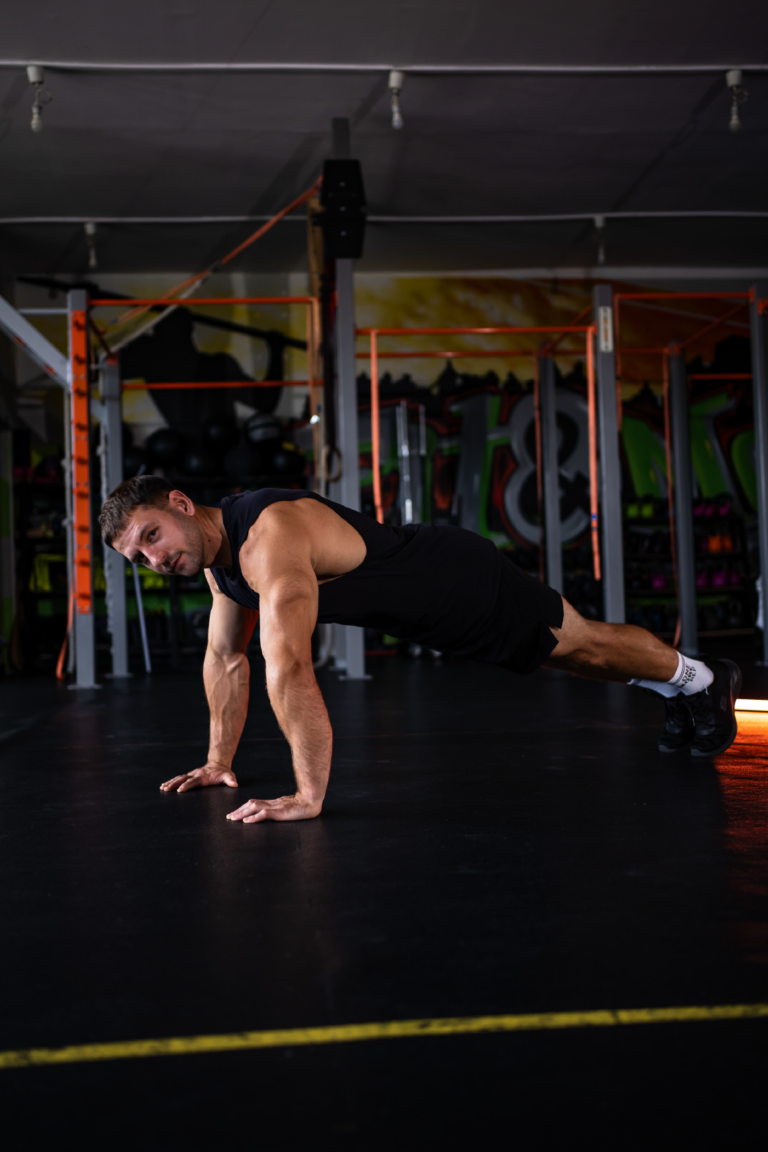 Man in a black sleeveless shirt performs a push-up on a gym floor with orange racks and graffiti walls in the background