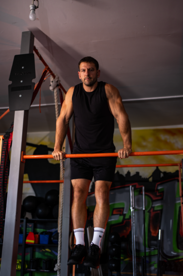 Man in a black sleeveless shirt and shorts doing a pull-up on an orange bar in a gym.