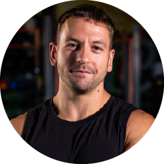 Portrait of a man with short dark hair and stubble, wearing a black sleeveless shirt, looking at the camera.