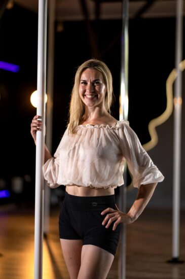 Smiling woman in a white, off-the-shoulder blouse and black shorts stands beside a pole in a dim studio, posing for the camera.