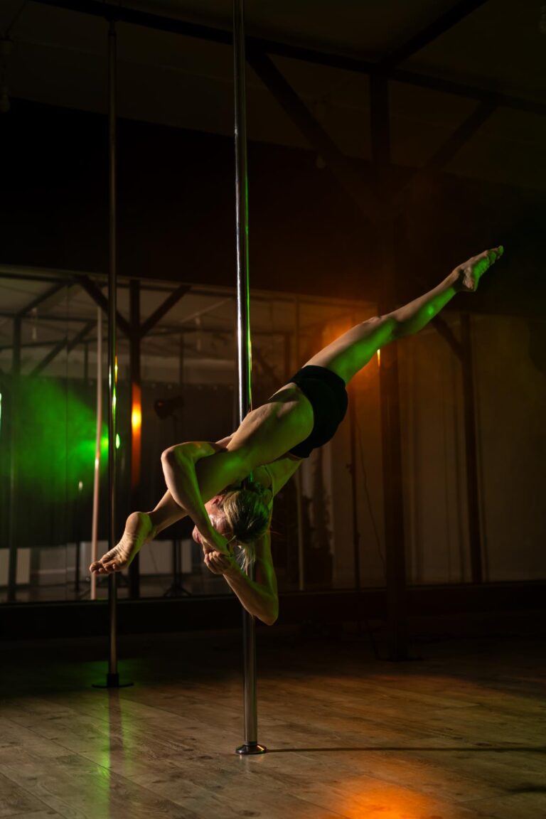 Pole dancer in a dim studio performs an inverted hold on a vertical pole with legs extended, green and orange lighting surrounding her.