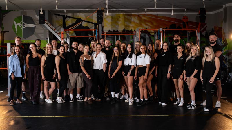 Large group photo of diverse people posing together in a gym with weight racks and colorful wall art in the background, facing the camera.